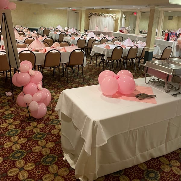 A banquet hall set up for an event, featuring tables with pink and white decor, and pink balloons.