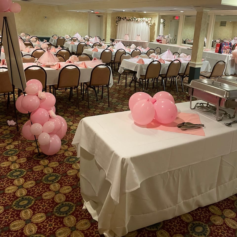 A banquet hall set up for an event, featuring tables with pink and white decor, and pink balloons.
