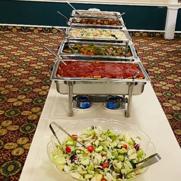 A buffet line with chafing dishes filled with pasta, vegetables, and other hot foods, plus a large salad bowl.