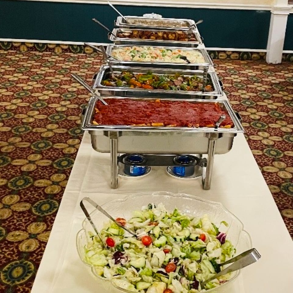 A buffet line with chafing dishes filled with pasta, vegetables, and other hot foods, plus a large salad bowl.